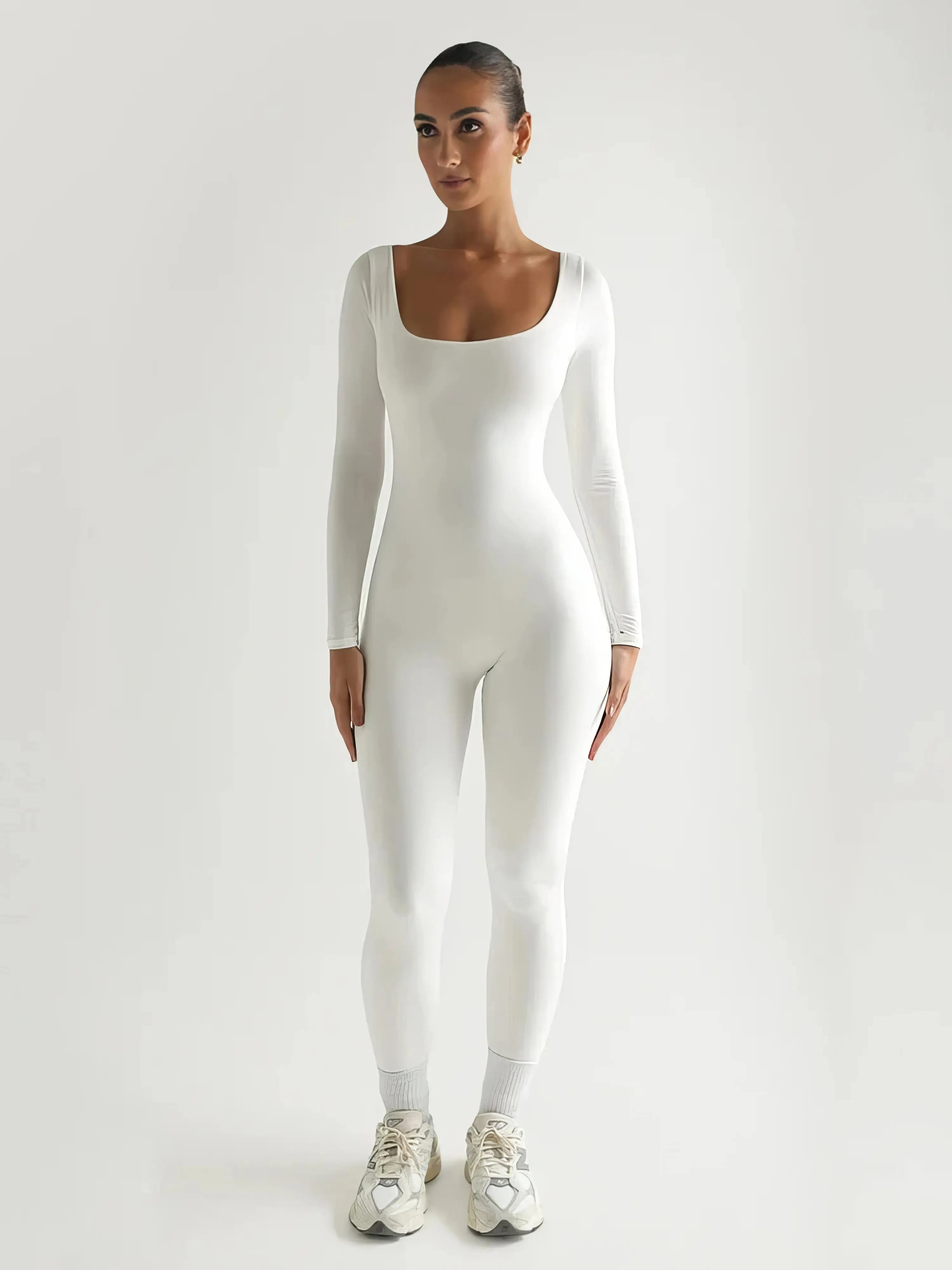 Tuta bianca aderente a maniche lunghe, indossata da una donna in piedi, scarpe da ginnastica bianche, sfondo neutro. Moda donna, abbigliamento sportivo.