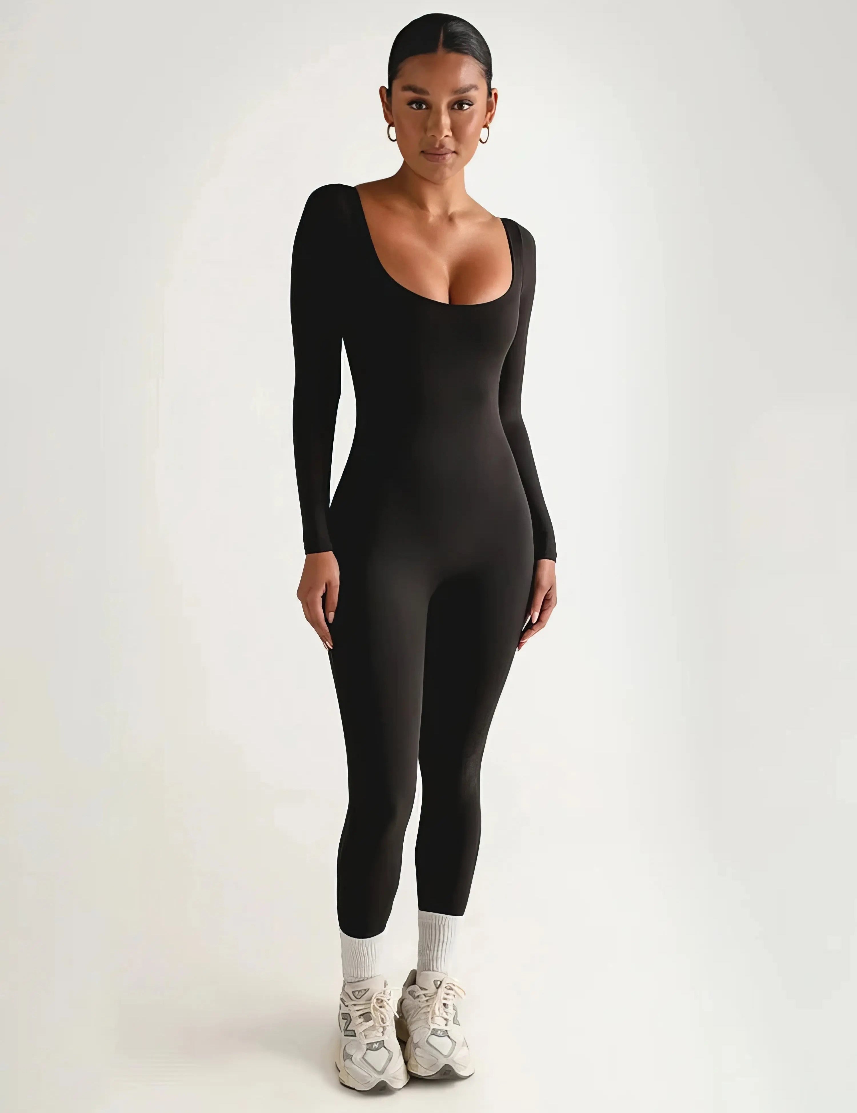 Donna in tuta nera aderente a maniche lunghe, scarpe da ginnastica bianche, sfondo neutro. Abbigliamento sportivo elegante, moda donna.