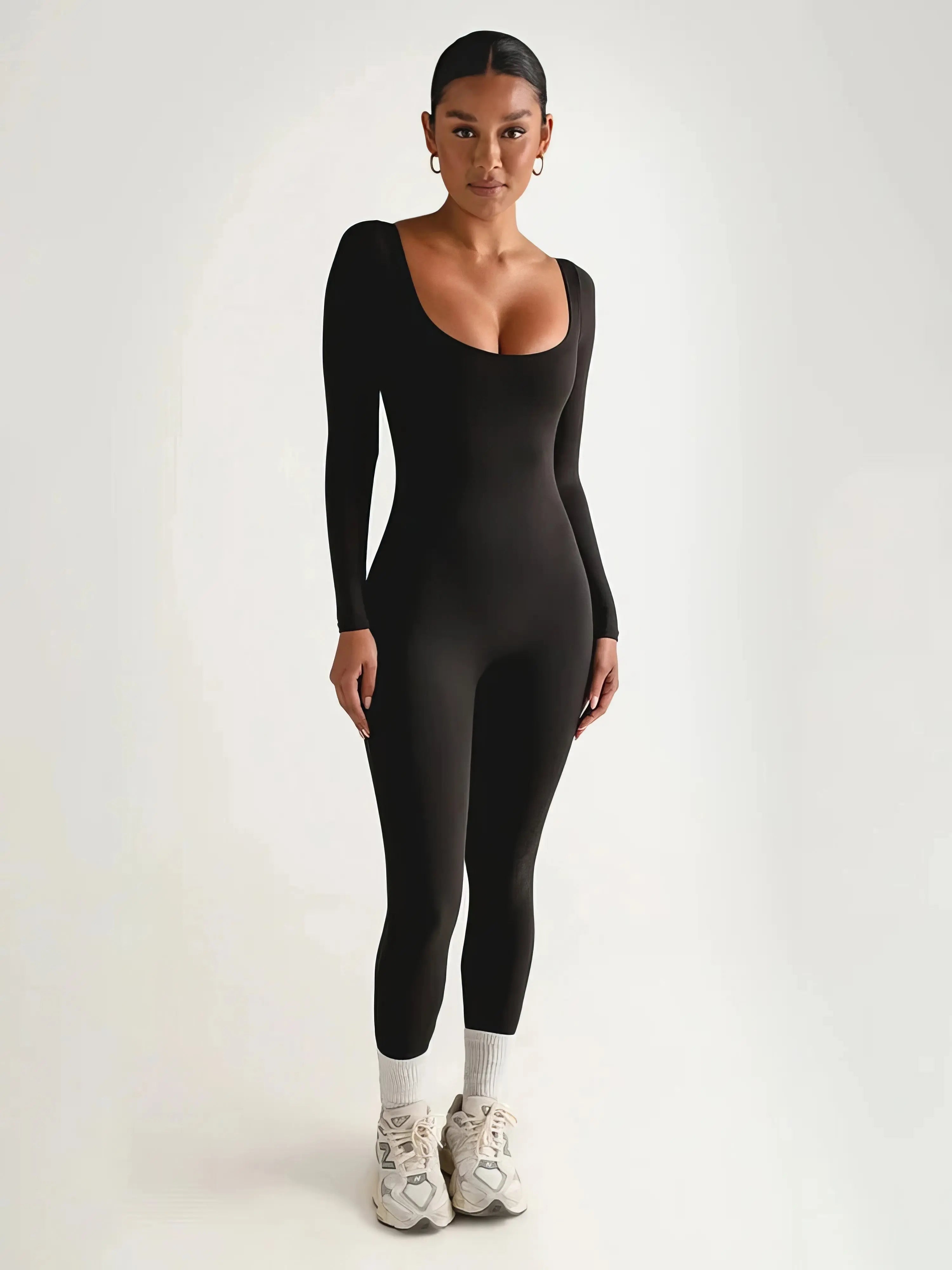 Donna in tuta nera aderente a maniche lunghe, scarpe da ginnastica bianche, sfondo neutro. Abbigliamento sportivo elegante, moda donna.