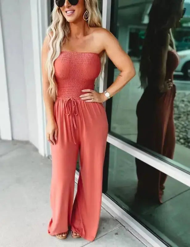 Donna con tuta intera rosa senza spalline, occhiali da sole e capelli lunghi ondulati, in piedi vicino a una vetrina. Moda estiva casual elegante.