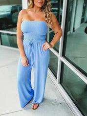 Donna con tuta jumpsuit azzurra senza spalline, tessuto arricciato, pantaloni larghi, sandali marroni, posa all'aperto vicino a vetrina.