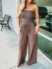 Donna con tuta intera marrone senza spalline, pantaloni larghi, davanti a una vetrina. Moda estiva, abbigliamento casual elegante.
