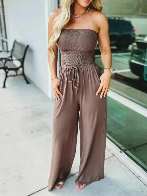 Donna con tuta intera marrone senza spalline, pantaloni larghi, davanti a una vetrina. Moda estiva, abbigliamento casual elegante.
