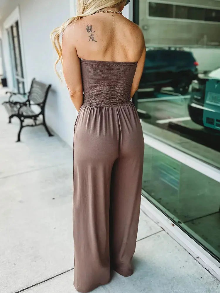 Donna in tuta marrone senza spalline, vista posteriore, tatuaggio sulla spalla, vicino a una vetrina. Moda estiva, abbigliamento casual elegante.