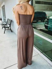 Donna in tuta marrone senza spalline, vista posteriore, tatuaggio sulla spalla, vicino a una vetrina. Moda estiva, abbigliamento casual elegante.
