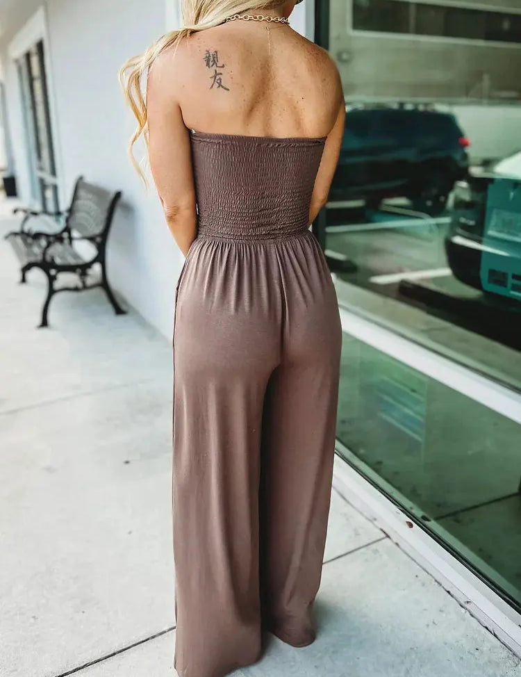 Donna in tuta marrone senza spalline, vista posteriore, tatuaggio sulla spalla, vicino a una vetrina. Moda estiva, abbigliamento casual elegante.