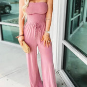 Donna in tuta rosa senza spalline, con borsa di paglia, davanti a una vetrina. Moda estiva, abbigliamento casual elegante, tuta femminile.
