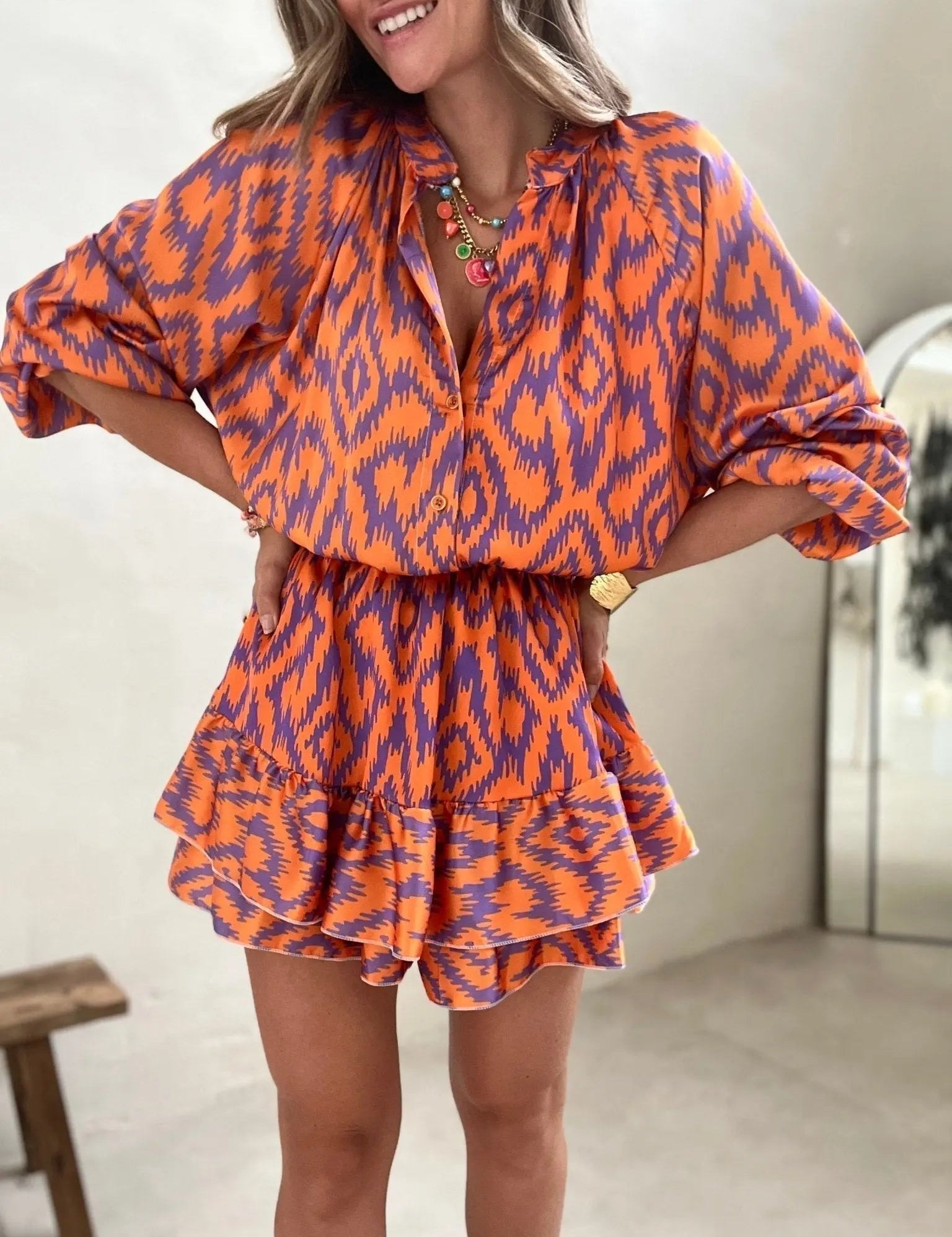 Abito corto donna arancione e viola con motivo geometrico, maniche lunghe, scollo a V, cintura in vita, collana dorata. Moda estiva elegante.