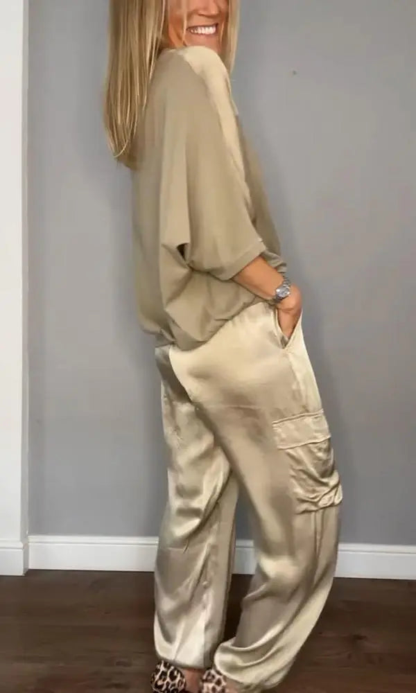 Donna in abbigliamento elegante beige, indossa pantaloni di seta e maglia morbida, posa sorridente su sfondo grigio. Moda femminile chic.