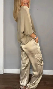 Donna in abbigliamento elegante beige, indossa pantaloni di seta e maglia morbida, posa sorridente su sfondo grigio. Moda femminile chic.