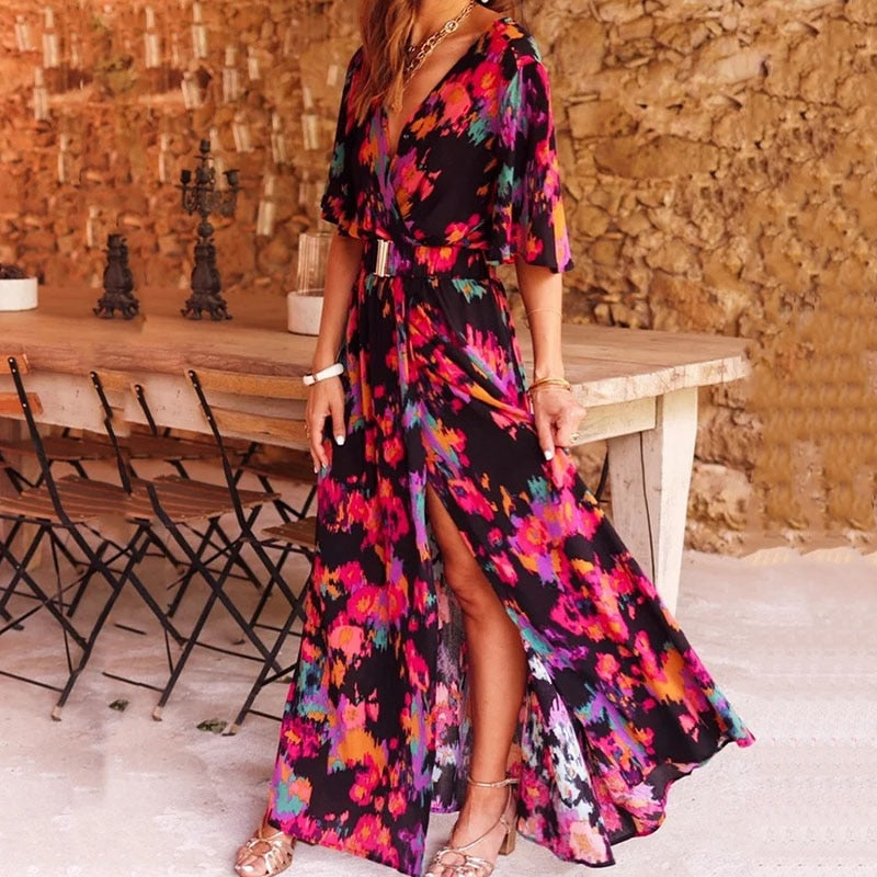 Abito lungo floreale colorato, donna, con spacco, maniche corte, sfondo rustico, tavolo in legno, sedie pieghevoli, moda estiva elegante.