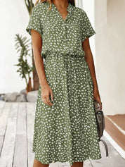 Abito estivo verde a pois bianchi, maniche corte, cintura in vita, perfetto per moda casual donna. Sfondo esterno con piante.