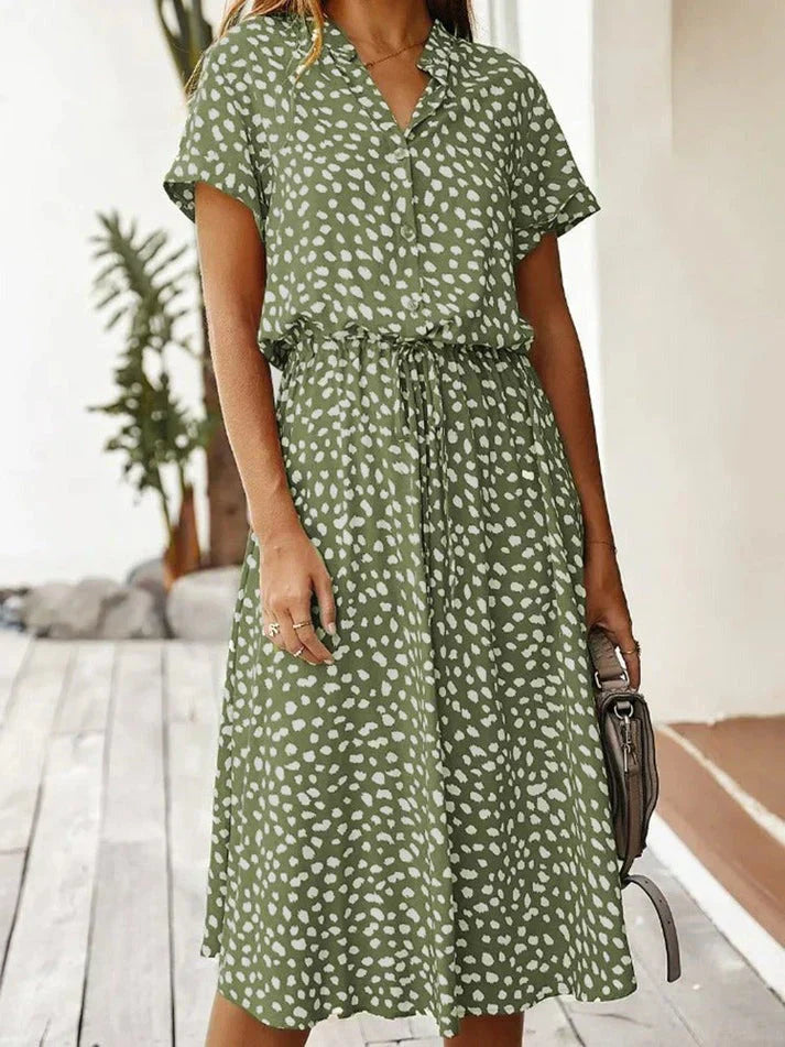 Abito estivo verde a pois bianchi, maniche corte, cintura in vita, perfetto per moda casual donna. Sfondo esterno con piante.