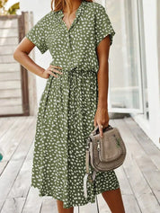 Abito estivo verde a pois bianchi, maniche corte, cintura in vita, donna in posa su terrazza. Borsa a tracolla beige. Moda casual femminile.