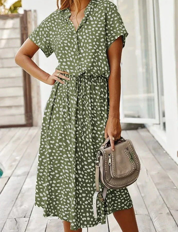 Abito estivo verde a pois bianchi, maniche corte, cintura in vita, donna in posa su terrazza. Borsa a tracolla beige. Moda casual femminile.