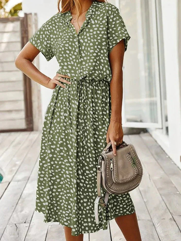 Abito estivo verde a pois bianchi, maniche corte, cintura in vita, donna in posa su terrazza. Borsa a tracolla beige. Moda casual femminile.