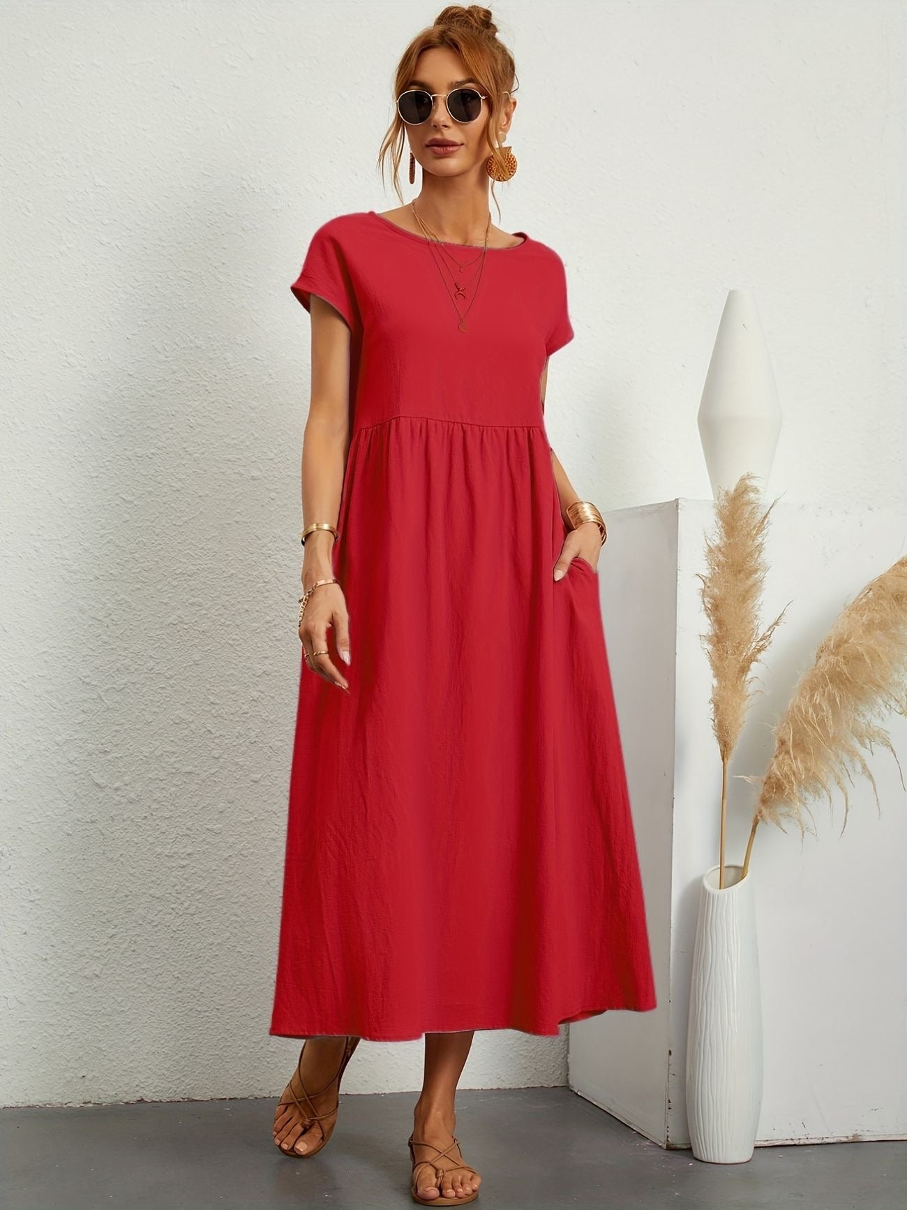 Donna con abito lungo rosso, occhiali da sole, sandali beige. Sfondo bianco minimalista, decorazione con pampas. Moda estiva elegante.