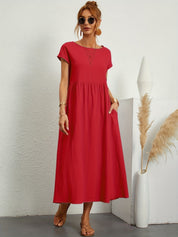 Donna con abito lungo rosso, occhiali da sole, sandali beige. Sfondo bianco minimalista, decorazione con pampas. Moda estiva elegante.