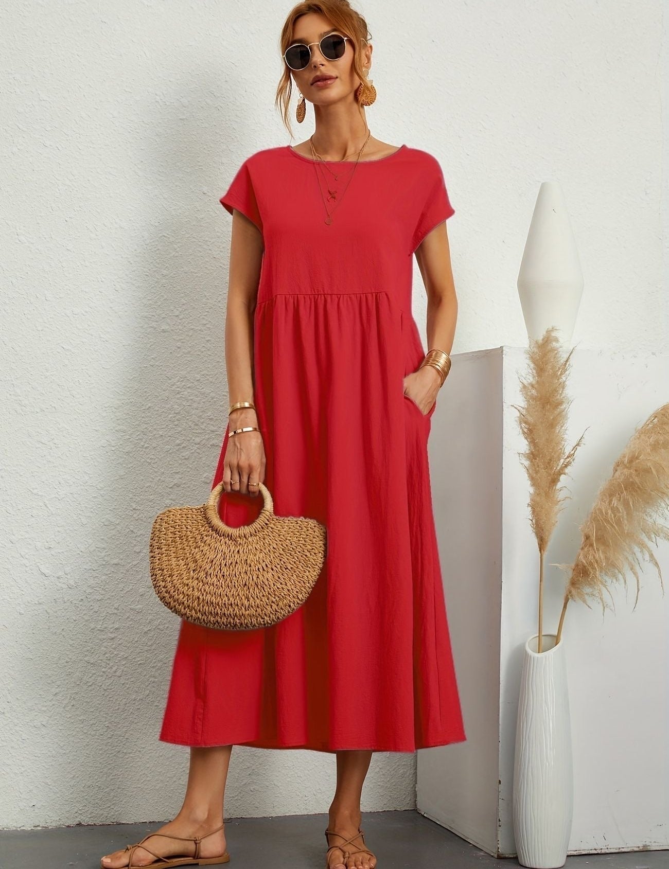 Donna con abito rosso estivo, borsa di paglia e sandali, posa accanto a vaso decorativo. Moda femminile casual, abbigliamento estivo elegante.