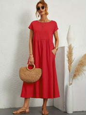 Donna con abito rosso estivo, borsa di paglia e sandali, posa accanto a vaso decorativo. Moda femminile casual, abbigliamento estivo elegante.