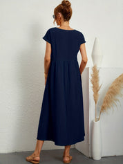 Donna con abito lungo blu, sandali beige, capelli raccolti, occhiali da sole, accanto a vaso bianco con pampas. Moda estiva elegante e casual.