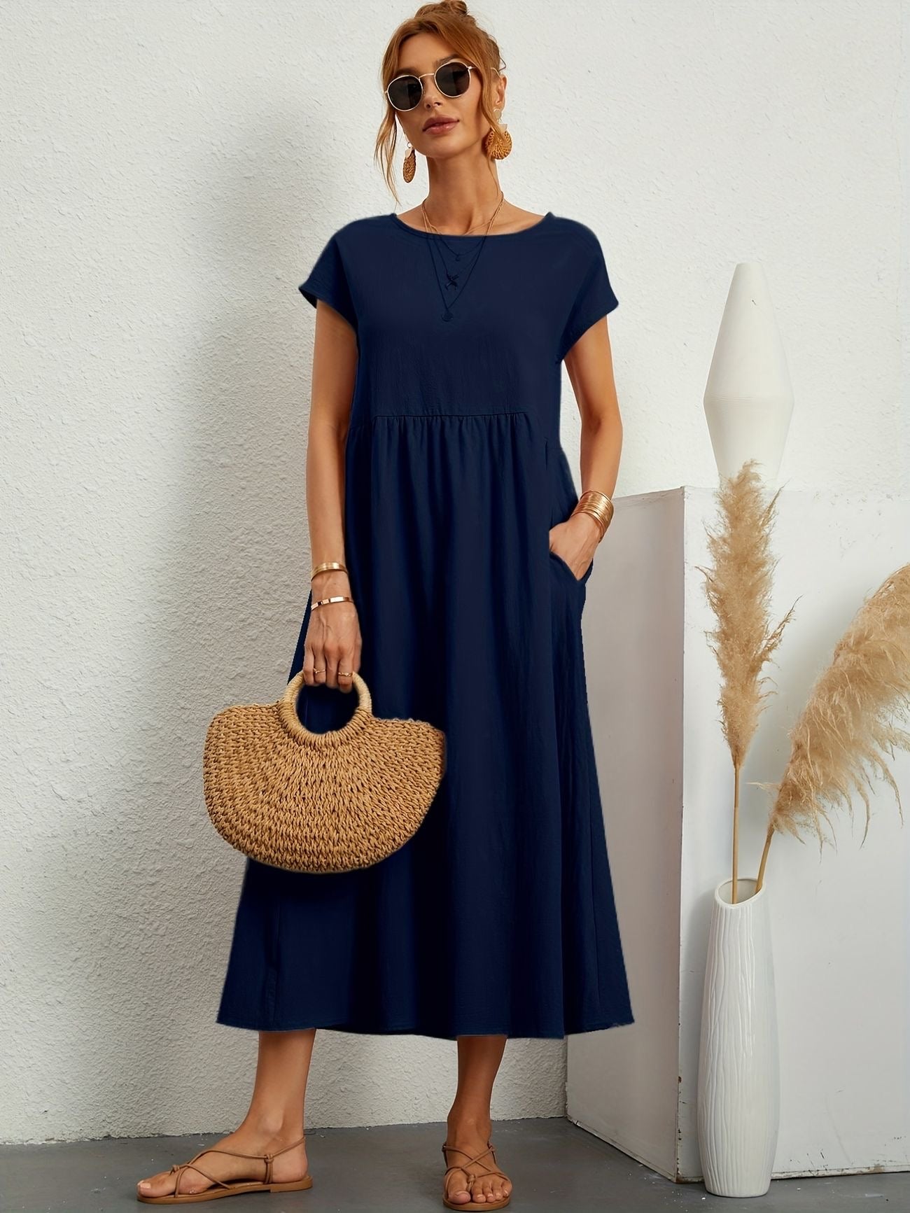 Donna con abito lungo blu navy, borsa di paglia, occhiali da sole e sandali beige. Sfondo bianco con decorazioni minimaliste. Moda estiva elegante.