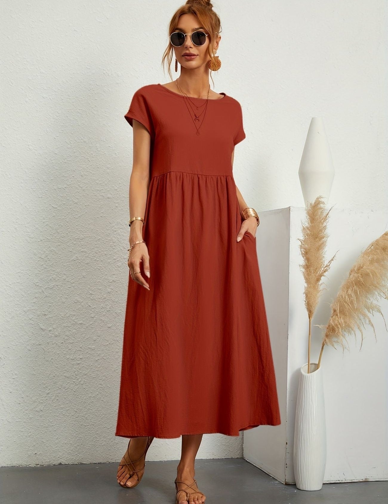 Donna con abito lungo rosso, occhiali da sole, sandali, accanto a vaso bianco con pampas. Moda estiva elegante, abbigliamento casual chic.