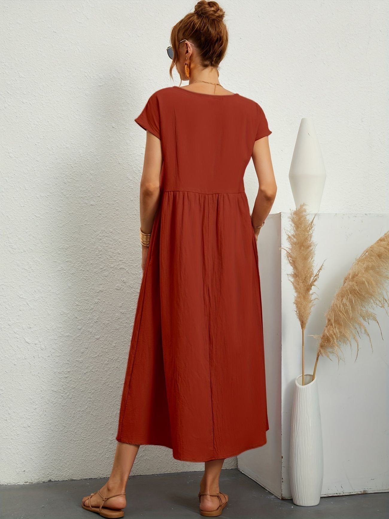 Donna con abito lungo rosso terracotta, sandali beige, vaso bianco con pampas. Moda estiva, abbigliamento casual elegante.