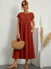 Donna con abito lungo rosso terracotta, borsa in paglia, occhiali da sole, sandali beige. Moda estiva elegante, stile casual chic.