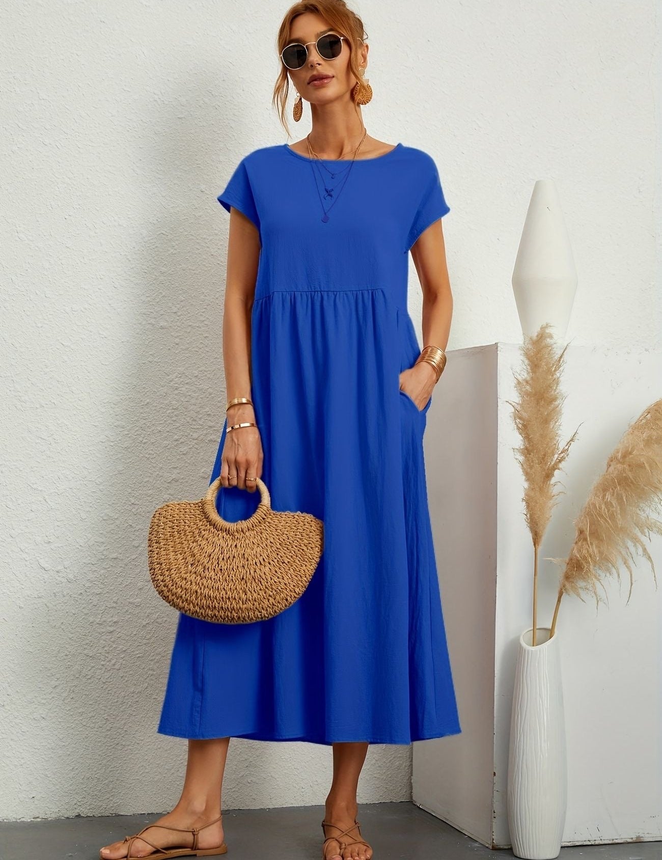 Donna con abito lungo blu, borsa di paglia e sandali, posa accanto a vaso decorativo. Moda estiva, abbigliamento casual elegante.