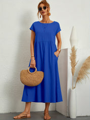 Donna con abito lungo blu, borsa di paglia e sandali, posa accanto a vaso decorativo. Moda estiva, abbigliamento casual elegante.