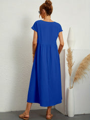 Donna con abito lungo blu, sandali beige, capelli raccolti, in piedi accanto a vaso bianco con pampas. Moda estiva, abbigliamento casual elegante.
