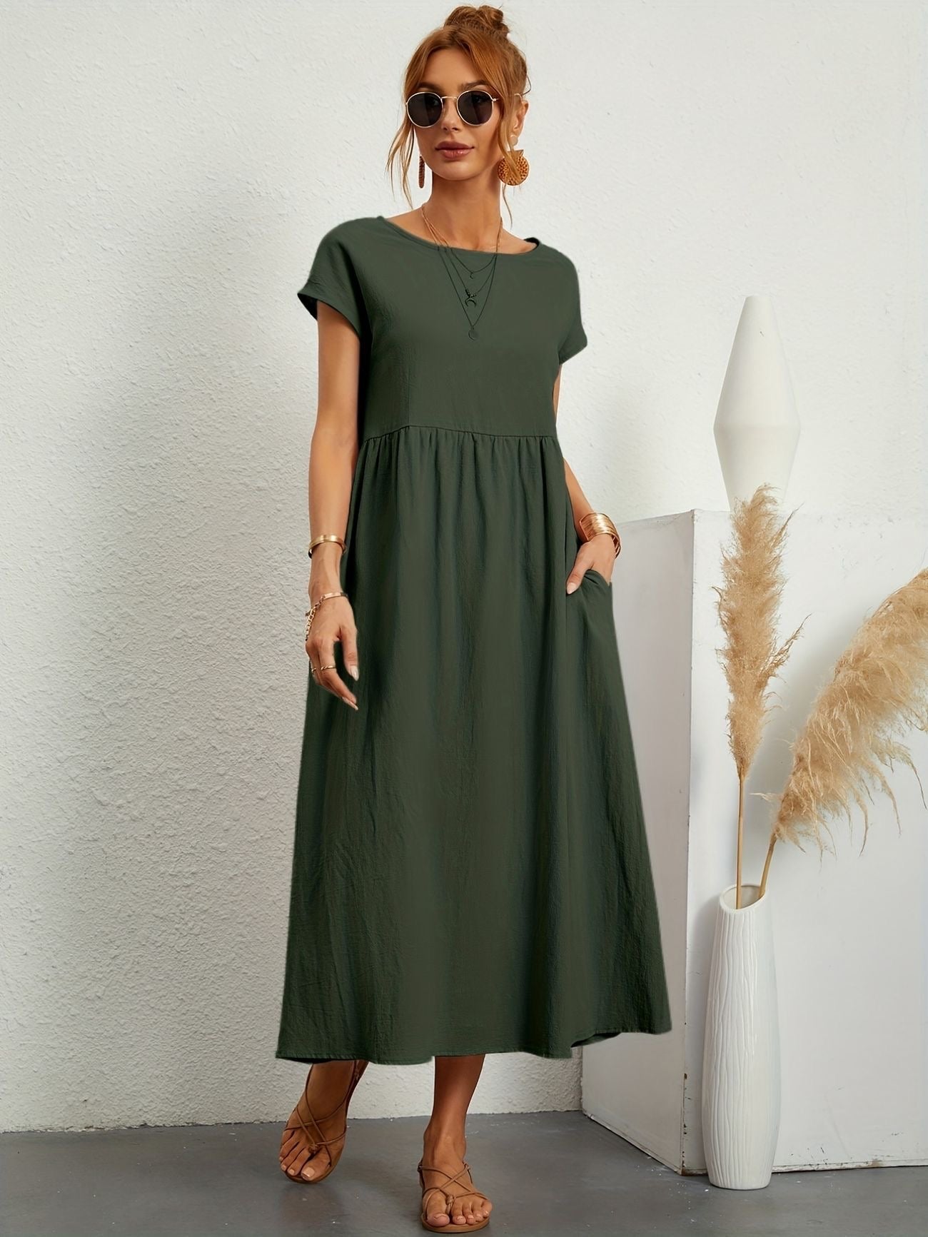 Donna con abito lungo verde oliva, occhiali da sole, sandali beige, accanto a vaso bianco con pampas. Moda estiva casual elegante.