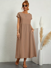 Donna con abito lungo marrone, occhiali da sole, sandali, accanto a vaso bianco con pampas. Moda estiva casual, abbigliamento femminile elegante.