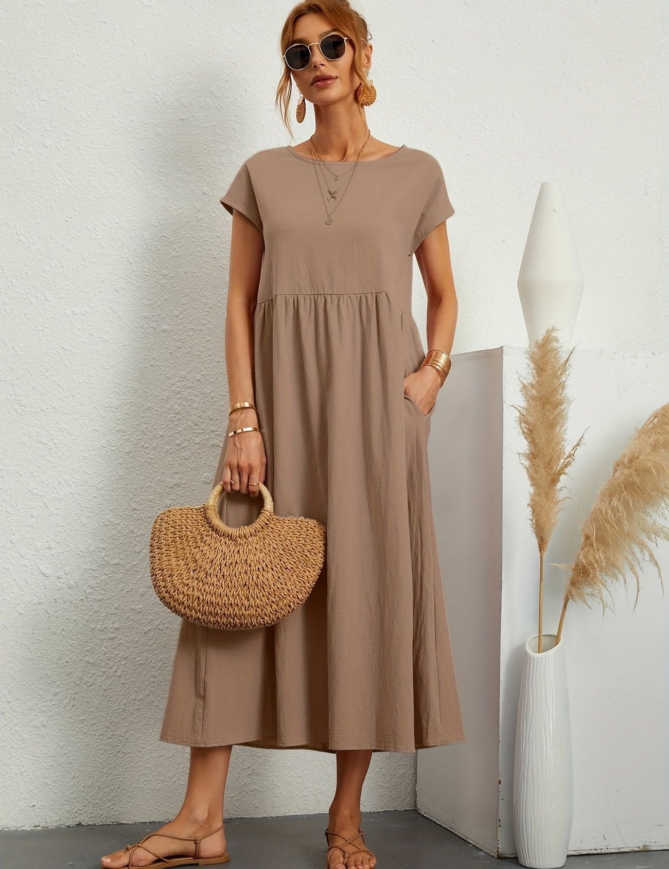 Donna con abito lungo beige, borsa di paglia, occhiali da sole, sandali, accanto a vaso bianco con pampas. Moda estiva elegante e casual.