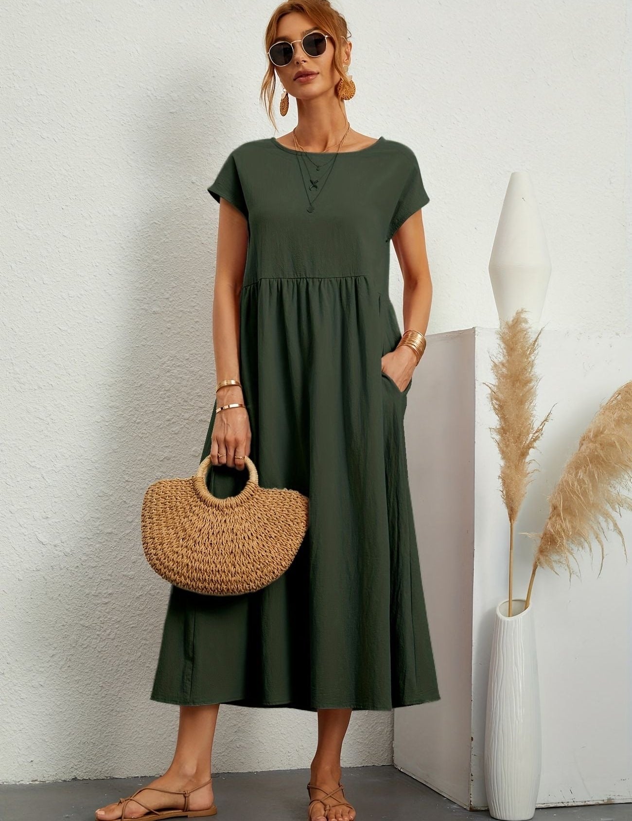 Donna con abito lungo verde, borsa di paglia e sandali, in posa accanto a vaso bianco con pampas. Moda estiva elegante e casual.