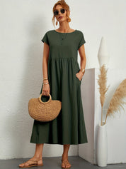 Donna con abito lungo verde, borsa di paglia e sandali, in posa accanto a vaso bianco con pampas. Moda estiva elegante e casual.