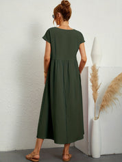 Donna con abito lungo verde oliva, sandali beige, accanto a vaso bianco con pampas. Moda estiva, abbigliamento casual elegante.