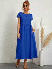 Donna con abito lungo blu, maniche corte, occhiali da sole, sandali marroni. Sfondo bianco con decorazioni minimaliste. Moda estiva elegante.