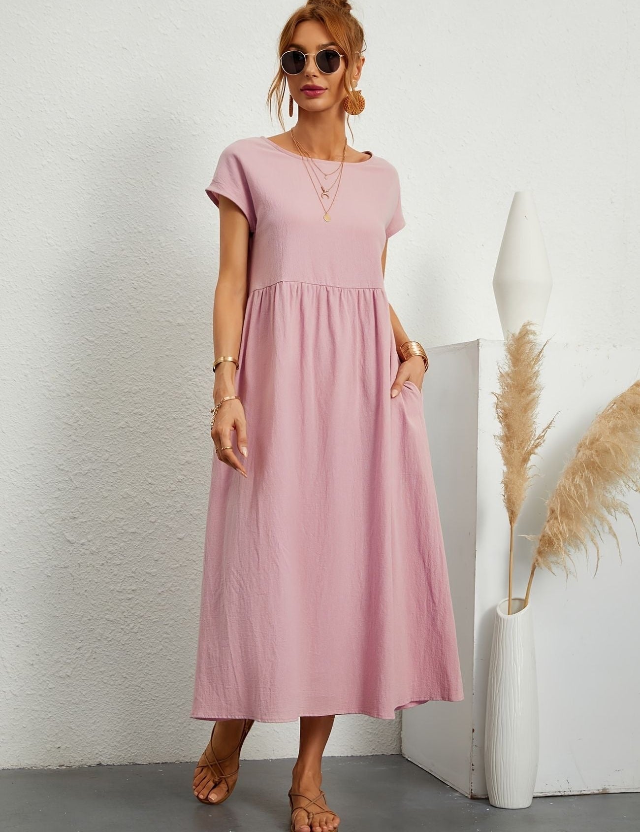 Donna con abito lungo rosa, occhiali da sole e sandali, accanto a vaso bianco con pampas. Moda estiva casual, abbigliamento femminile elegante.