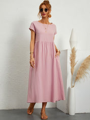 Donna con abito lungo rosa, occhiali da sole e sandali, accanto a vaso bianco con pampas. Moda estiva casual, abbigliamento femminile elegante.
