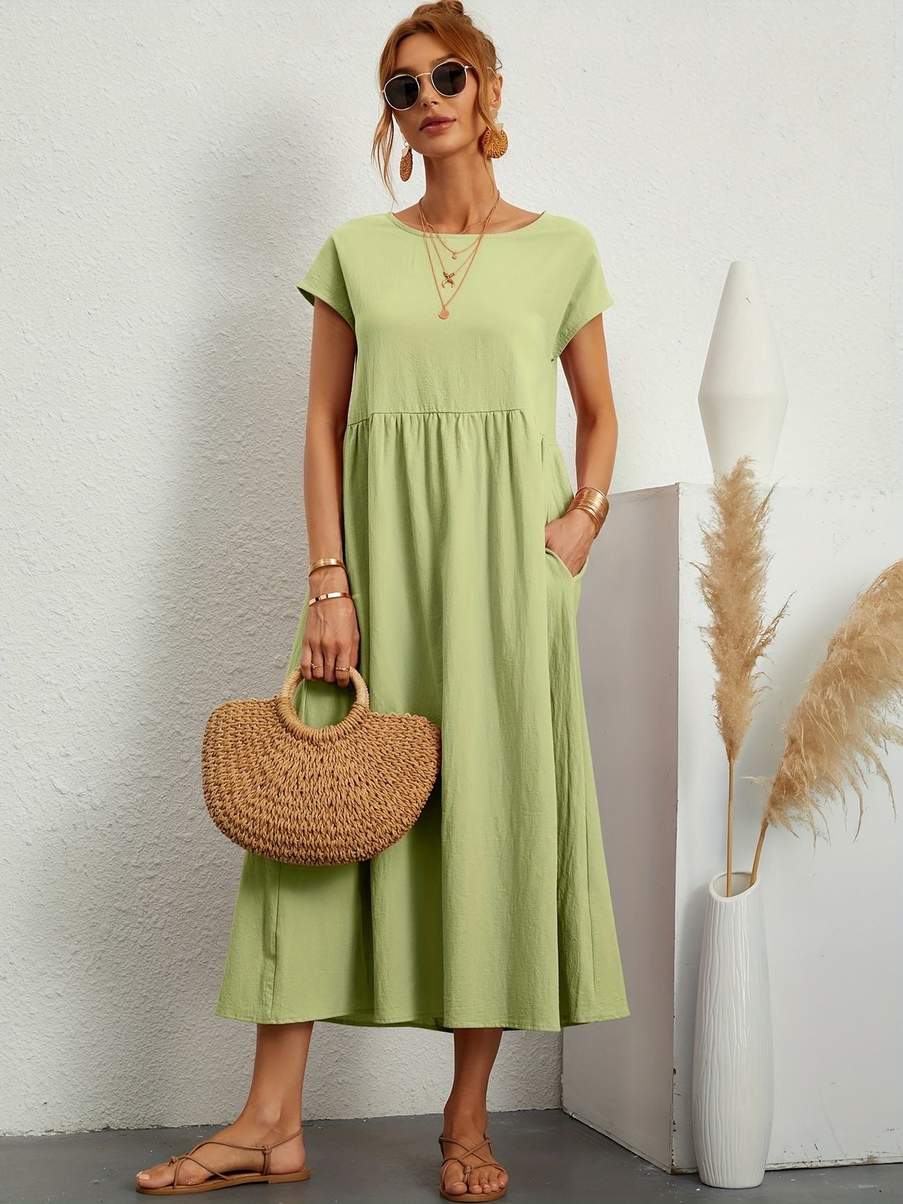Donna con abito lungo verde chiaro, borsa di paglia, sandali beige, occhiali da sole. Sfondo bianco minimalista, decorazione con pampa.
