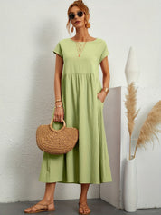 Donna con abito lungo verde chiaro, borsa di paglia, sandali beige, occhiali da sole. Sfondo bianco minimalista, decorazione con pampa.
