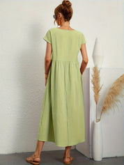 Donna con abito lungo verde chiaro, sandali beige, accanto a vaso bianco con pampas. Moda estiva, abbigliamento casual, stile minimalista.