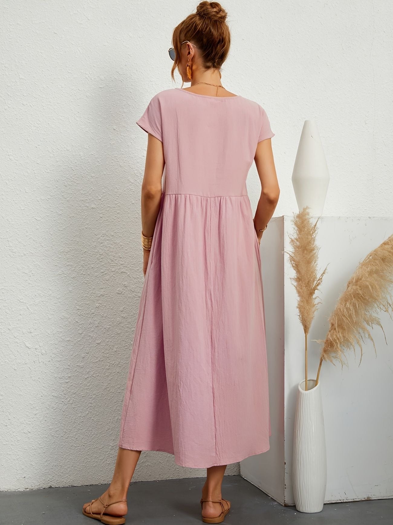 Donna con abito lungo rosa, sandali beige, in piedi accanto a vaso bianco con pampas. Moda estiva, abbigliamento casual elegante.