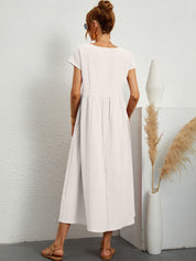 Donna di spalle con abito lungo bianco, sandali beige, accanto a vaso decorativo con pampas. Moda estiva, abbigliamento casual elegante.