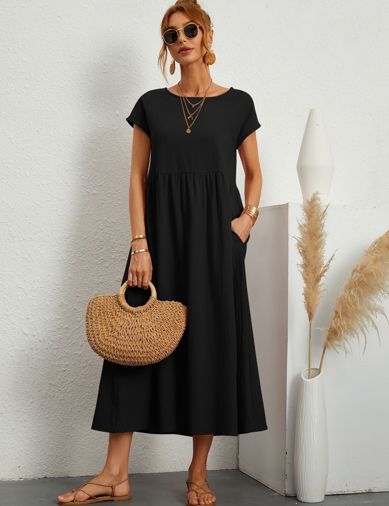 Donna con abito lungo nero, borsa di paglia e sandali, posa accanto a vaso decorativo. Moda estiva elegante, accessori oro, occhiali da sole.