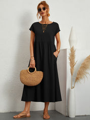 Donna con abito lungo nero, borsa di paglia e sandali, posa accanto a vaso decorativo. Moda estiva elegante, accessori oro, occhiali da sole.