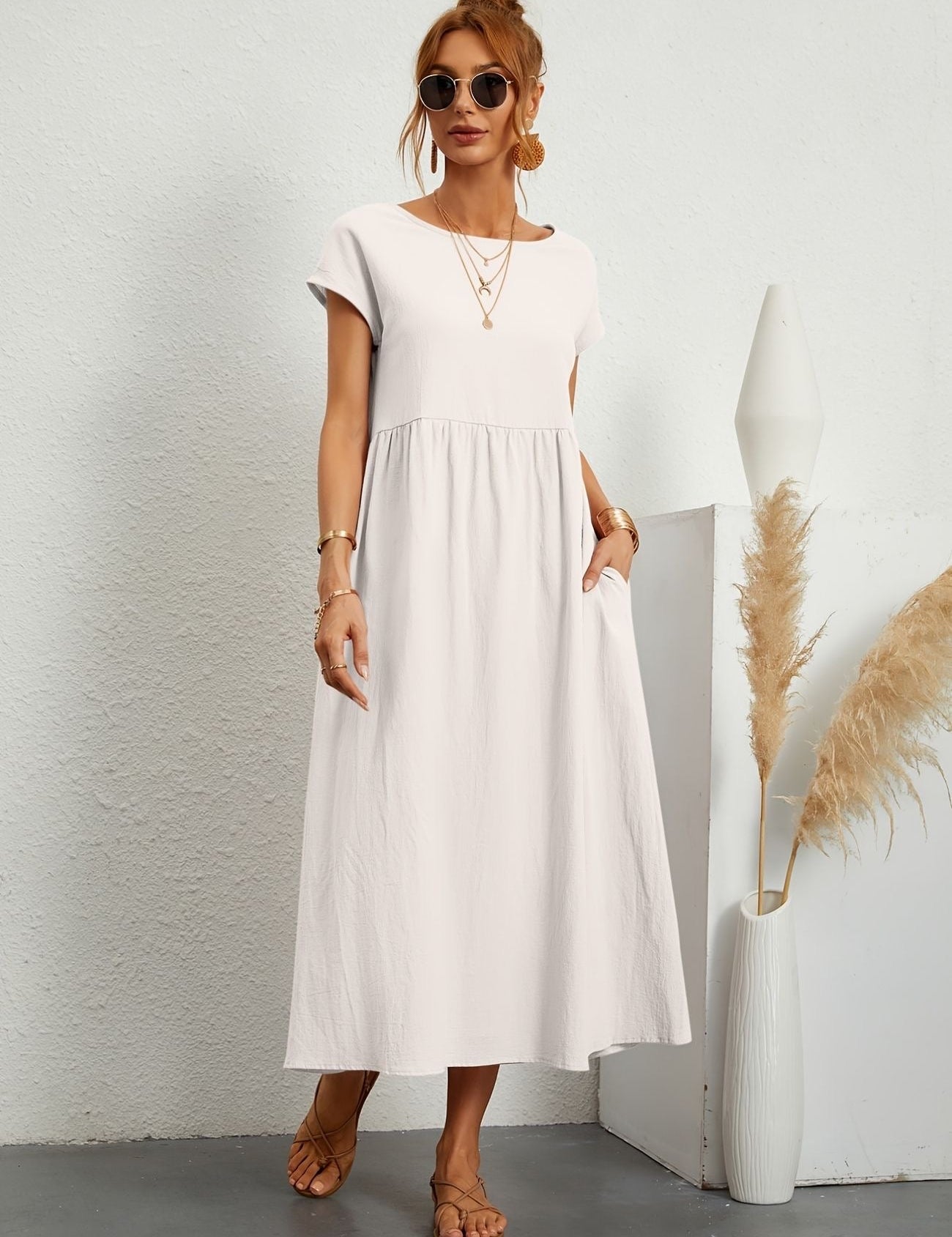 Donna con abito lungo bianco, occhiali da sole e sandali, accanto a vaso decorativo. Moda estiva elegante, abbigliamento casual chic.