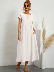 Donna con abito lungo bianco, occhiali da sole e sandali, accanto a vaso decorativo. Moda estiva elegante, abbigliamento casual chic.